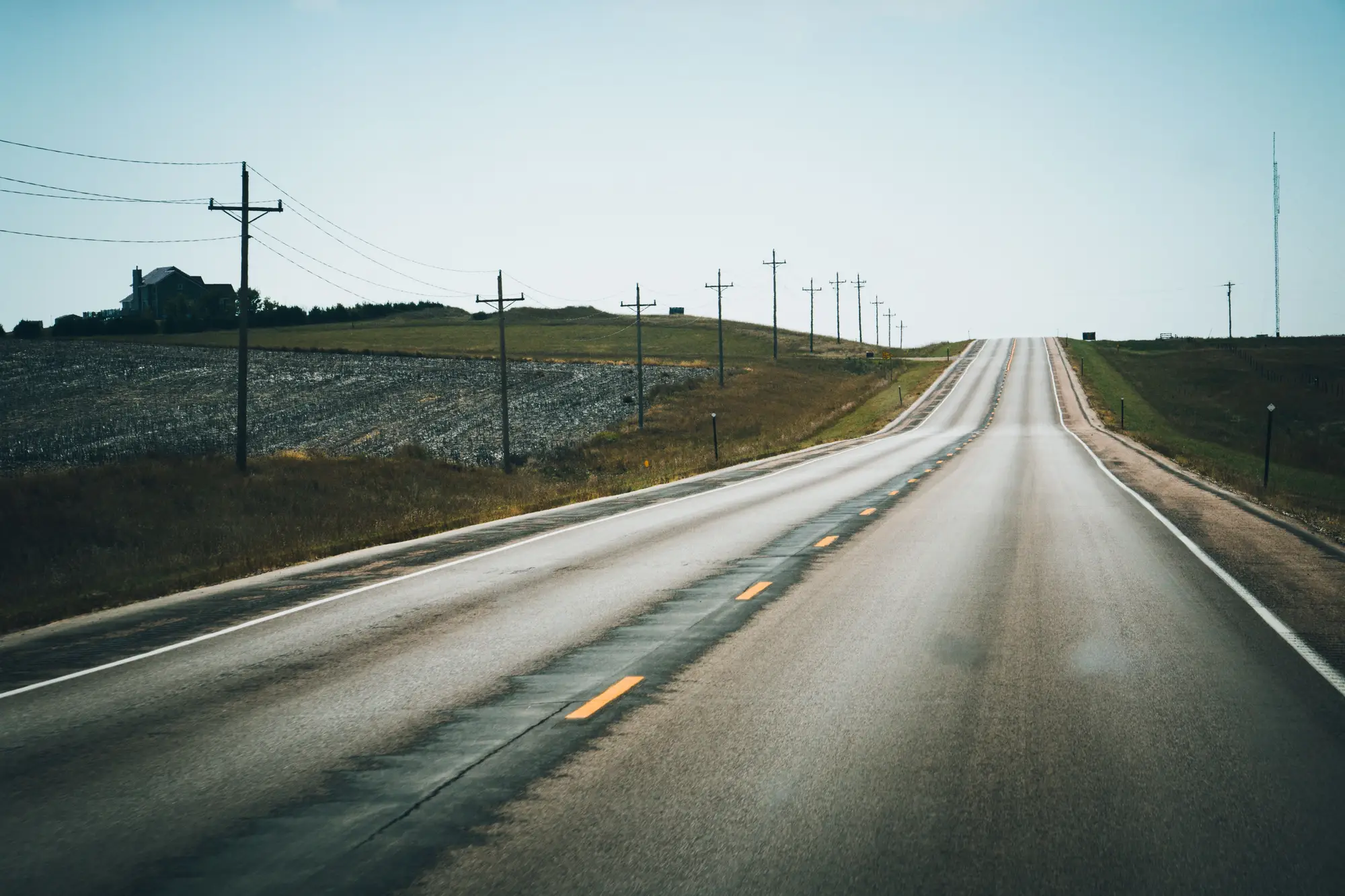 A two-lane rural highway running over a rise between fields and power poles, its centerline joint sealed and darkened